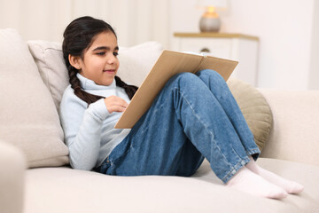 Fototapeta premium Cute little girl reading book on couch at home