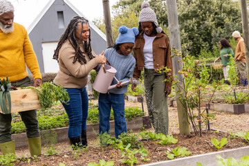 African American family gardening in garden mom with braids guiding child pouring pink watering can