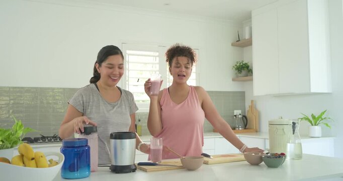 Diverse female pals lifting blender lid, making smoothie sipping from glasses and testing at island
