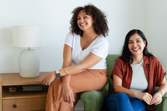 Two female friends sitting on green couch at home, wearing rust clothing and showing smartwatch