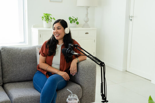 Woman sitting on gray sofa wearing white headphones and speaking into black microphone at home