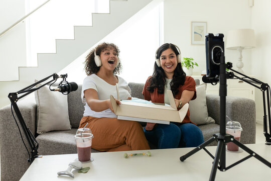 Diverse female friends recording with mics and opening product box on gray sofa in living room