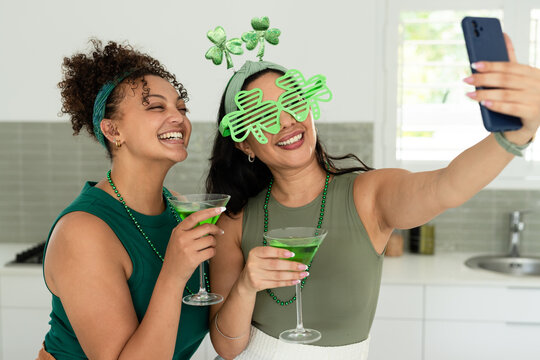 Diverse female friends posing in kitchen with shamrock glasses, holding green drinks and smartphone