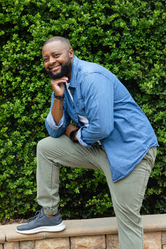African American man kneeling on stone retaining ledge by green hedge in garden, leaning, smiling