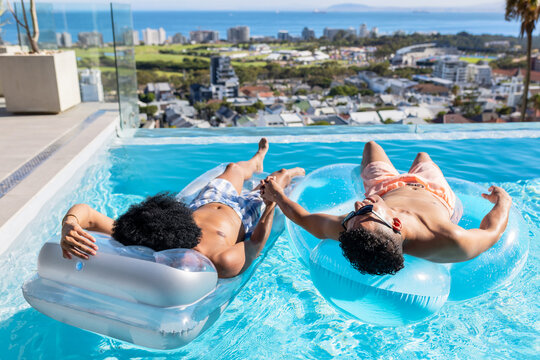 African men reclining on clear inflatable lounge and tube, holding hands on rooftop infinity pool