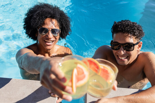 African American men wearing sunglasses clinking grapefruit cocktails leaning on blue pool edge