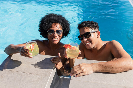 African American male friends shirtless leaning on pool edge holding citrus drinks, wearing shades