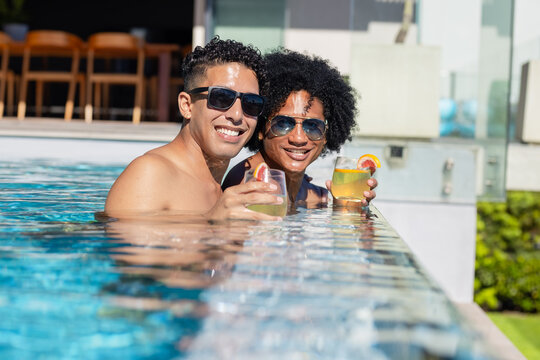 African American men standing in rooftop pool edge, holding cocktails in swim trunks and sunglasses