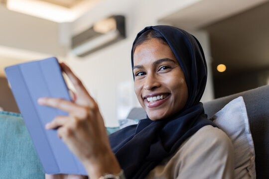 Woman wearing dark navy hijab reclining on gray couch at home holding blue tablet smiling