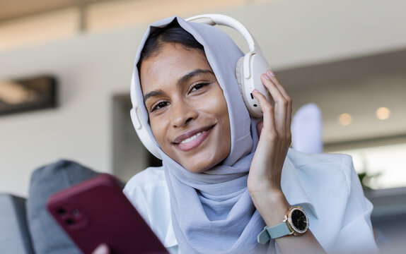 Woman wearing gray hijab and white headphones, holding red smartphone, showing mint watch on couch