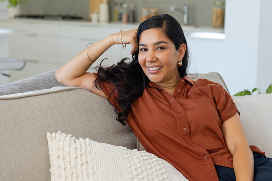 Middle-aged woman reclining on light gray sofa, wearing rust shirt and gold hoops, kitchen backdrop