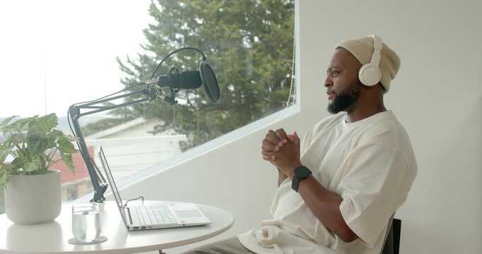 African man joining laptop recording call, speaking into mic at table by slanted window, copy space
