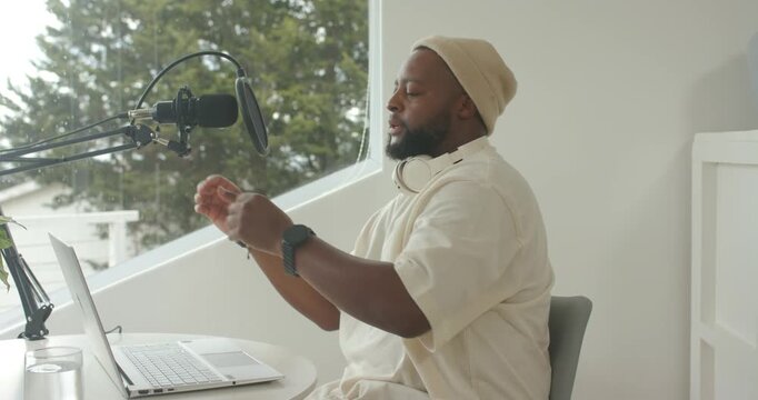 Adult African man reaching adjusting mic pop-filter at laptop donning headphones prepping to record