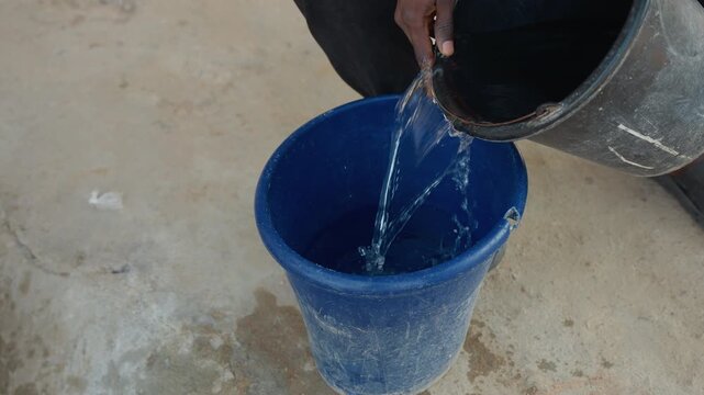 Blue bucket black pail house chores worker pours water from black pail into blue bucket on concrete floor visible hands repeated pouring sequence small splashes and drips outdoor courtyard cleaning