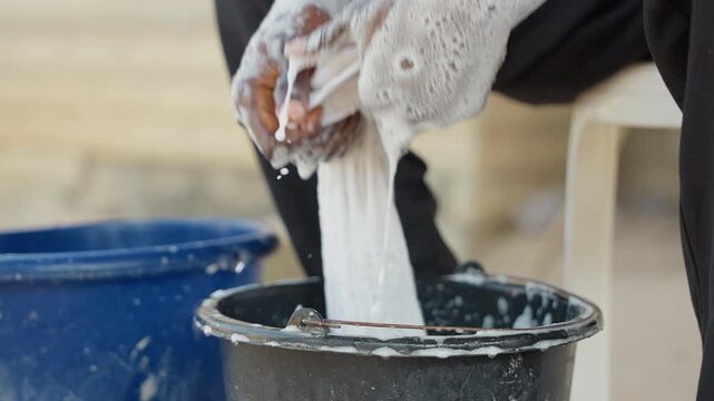 Black man wringing soapy cloth by house bucket on porch, sudsy hands, rhythmic motion, laundry worker energy, outdoor domestic scene, towel stretching and rinsing, focused routine