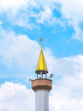 A mosque minaret with a golden spire topped by a crescent moon and star symbol, equipped with loudspeakers against a bright blue cloudy sky.