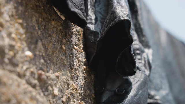 Black garment drying on stone wall crevice, closeup of damp sleeve tucked into rough mortar, gritty pebble detail and weathered texture, domestic laundry chore captured in muted light