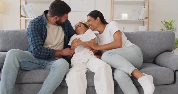 Happy family tickling daughter together at home. Parents on a couch as mother leans and father gives a smile; the girl giggles, filling the room with laughter. Concept: family fun at home.