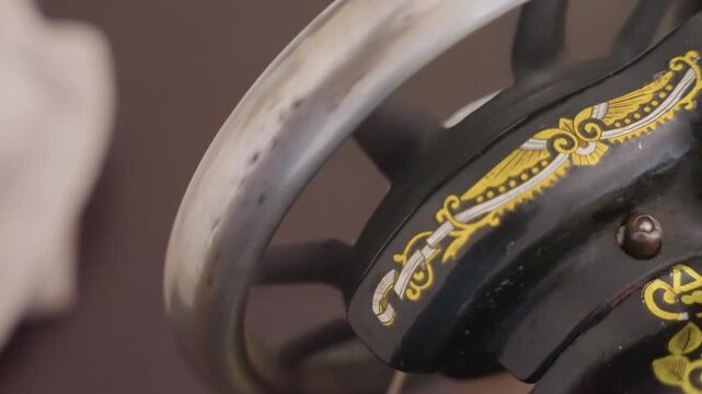 Black vintage sewing machine wheel closeup showing ornate gold decal and worn metal rim, intimate domestic repair scene in house workroom, hands on mending and restoration mood, slow crafty