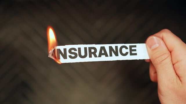 Close-up of a hand holding a paper strip labeled &ldquo;INSURANCE&rdquo; as the edge burns with flame on a dark background. Rising premiums and coverage crisis concept with copy space.