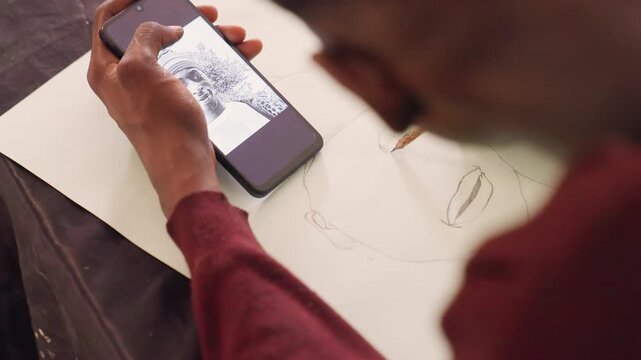 Artist sketching portrait with phone, pencil and paper. Overshoulder closeup of hands tracing soft outline while smartphone displays black painting reference, warm studio light, graphite lines,