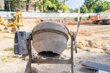 Portable concrete mixer with a weathered drum and metal frame stands on a dusty construction site, with excavators, earth piles and safety fencing in the background under bright midday sunlight
