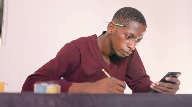 Black male artist painting with phone reference, balancing quick sketch and color notes at desk, intent posture in maroon sweater, indoor table scene with paint pots, determined productive mood