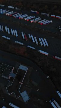 Vertical Screen Aerial Truck Parking Lot At Dusk Showing Rows Of Trailers Lined In Depot, Drone Perspective Captures Industrial Buildings, Long Queues Of Freight Trailers, Moody Low Light