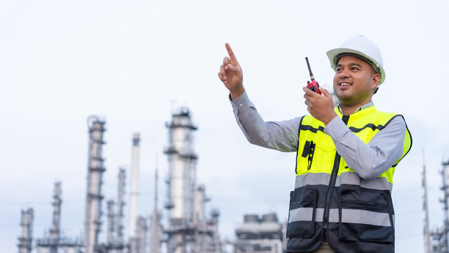 Happy handsome Asian man petrochemical engineer using walkie talkie with petroleum oil refinery in industrial estate background. Engineers at industry manufacturing.