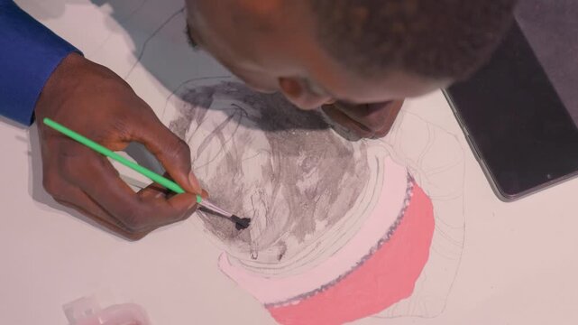 Overhead black artist painting black portrait with green brush, focused illustrator leaning over paper, careful tonal washes and charcoal lines build expressive face, studio table with phone