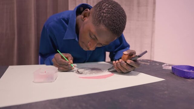 Black student painting at table focused on small brushwork, smartphone at hand, white paper on dark tabletop, palette cup and paint pot nearby, warm curtain backdrop, soft studio light
