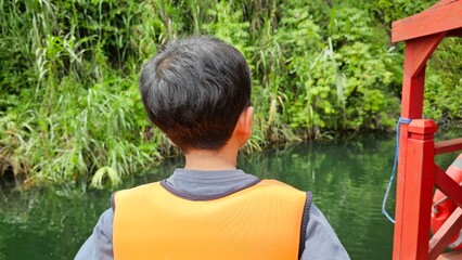 A boys sit on a boat wearing life vest and look at the view around the lake.