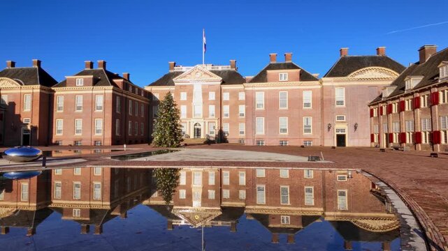 Low angle view of Paleis Het Loo main building reflected in still water during winter. Elegant Dutch Baroque architecture with festive Christmas tree in Apeldoorn museum grounds at midday.