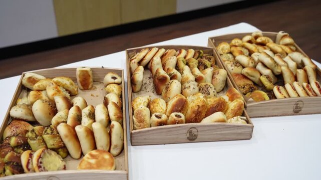 Wide shot of assorted breadsticks and mini pastries arranged in wooden trays on a buffet table indoors, showcasing bakery catering selection under bright lighting.