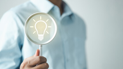 Business professional holding magnifying glass with light bulb icon, symbolizing idea research, innovation discovery, creative thinking and strategic business analysis concept. © Garun Studios