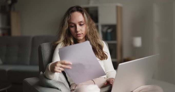 Young adult woman sits in armchair with laptop, hold papers, reviewing university admission documents, analyze paperwork, checking bank statements, reading official correspondence at home. Remote work