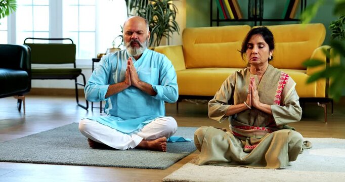 Indian senior couple doing omkar pranayama or yoga at home in the morning sitting with folded legs in modern living room chanting aum, practicing mindful breathing exercise for health