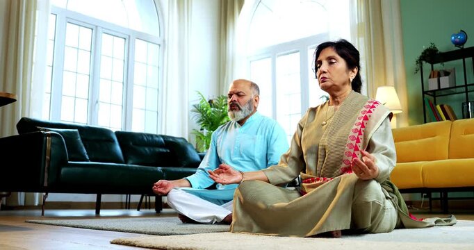 Indian senior couple doing omkar pranayama or yoga at home in the morning sitting with folded legs in modern living room chanting aum, practicing mindful breathing exercise for health