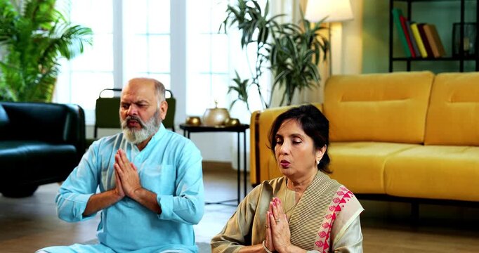 Indian senior couple doing omkar pranayama or yoga at home in the morning sitting with folded legs in modern living room chanting aum, practicing mindful breathing exercise for health