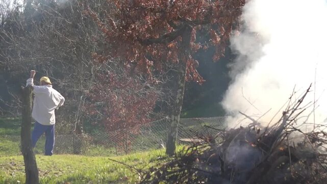 An elderly man stands by a wooden post, observing a large pile of dry branches burning in a grassy field, showcasing a traditional rural lifestyle and garden maintenance.