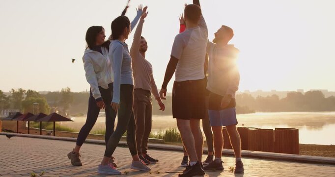 Motivated group of young active runners putting hands together and celebrating successful outdoor training. Happy jogging team standing in circle, showing team spirit, unity and support. Slow motion.