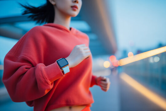 East Asian Female Runner In Red Hoodie With Smartwatch, Urban Evening Jogging And Fitness Tracking Concept