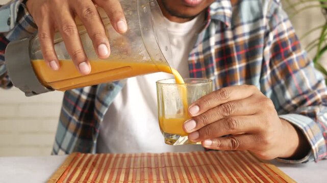 Young man pouring fresh orange mango smoothie from blender into glass on table