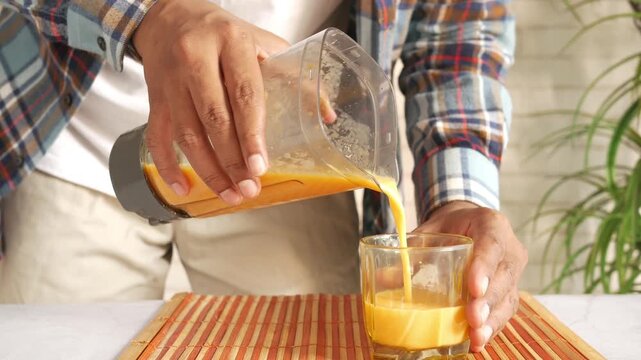 A man pouring fresh orange juice from a blender into a clear glass on a wooden mat, capturing the dynamic motion of liquid