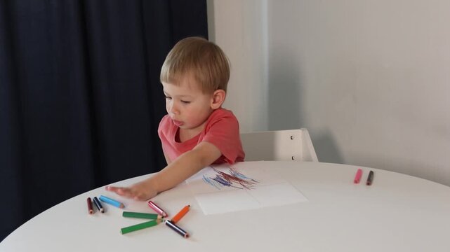 A preschool-aged boy draws lines with a pencil, making strokes on a white sheet of paper. The boy scribbles on the paper.