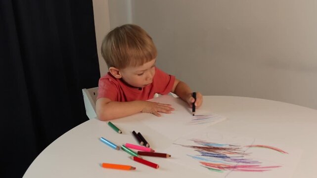 A preschool-aged boy draws lines with a pencil, making strokes on a white sheet of paper. The boy scribbles on the paper