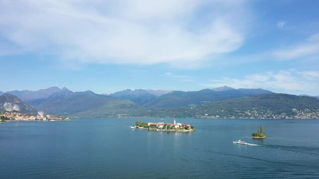 A scenic view of Isola dei Pescatori on Lake Maggiore, Italy, with surrounding mountains and a boat on the water under a blue sky.