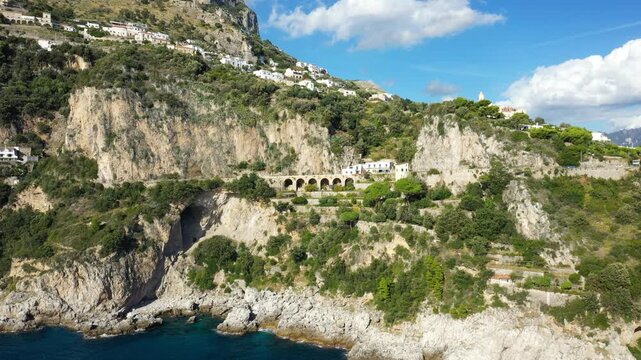 Aerial view of rugged cliffs and hillside villas overlooking the turquoise sea on Capo di Conca, Amalfi Coast, Italy. Lush greenery and dramatic rock formations highlight the Mediterranean landscape.