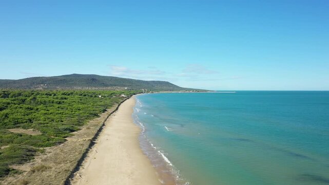 Drone shot of a pristine sandy beach and turquoise sea along the Isola Varano natural reserve, with lush greenery and distant hills under a clear blue sky.