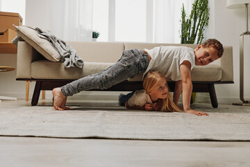 Two children playfully exercise indoors, a boy in a plank pose on the back of a girl lying on the...